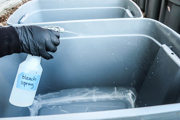 A gloved hand spraying a plastic tote with a bottle labeled 