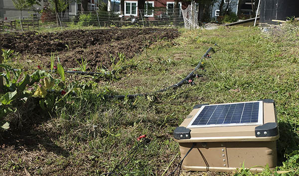 A solar energizer placed outside in the sun, in the background are crop rows where vegetables grow.