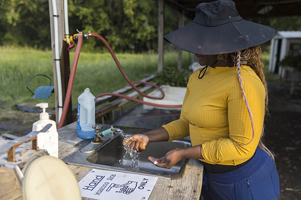 a black skinned farmer wearing a yellow shirt and black hat that covers her face is washing her hands at an infield wash station.  The water is running over her right hand from the spigot