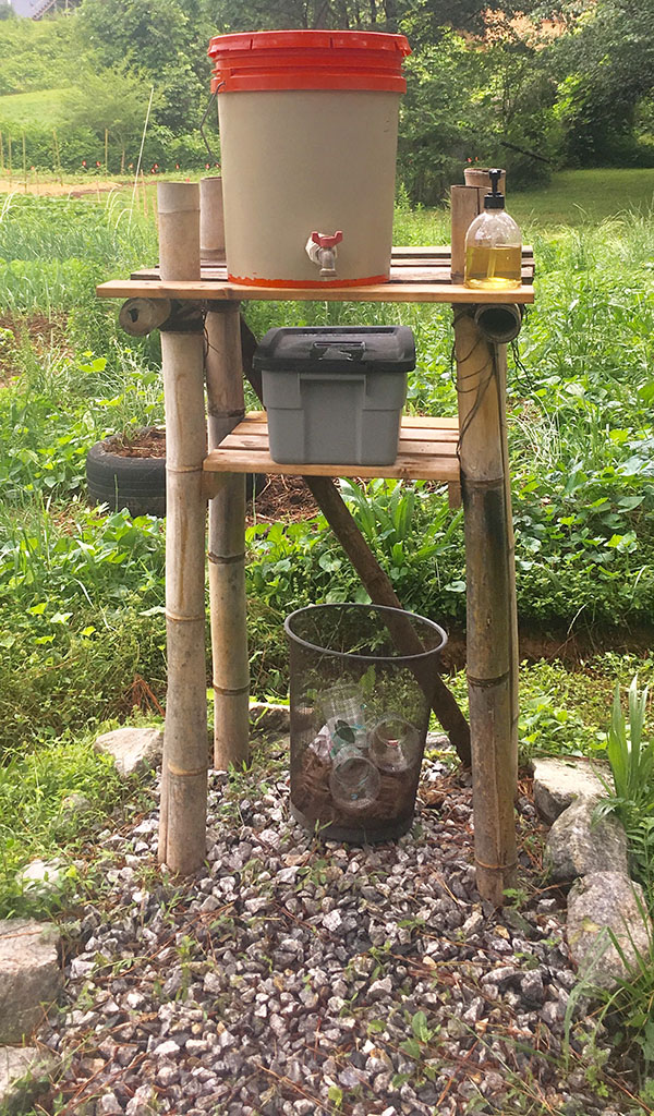 A portable handwash station. A five-gallon bucket with a spigot at the bottom sits on top of a stand made from bamboo. The stand also contains liquid hand soap in a bottle, a box of paper towels, and a wastebasket.