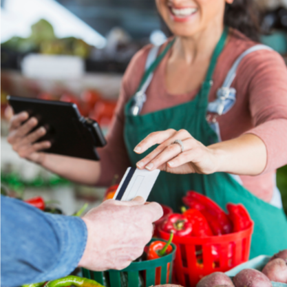 A customer hands a credit card to a vendor across a farmers' market table. The vendor is wearing a green apron and displaying red peppers for sale.